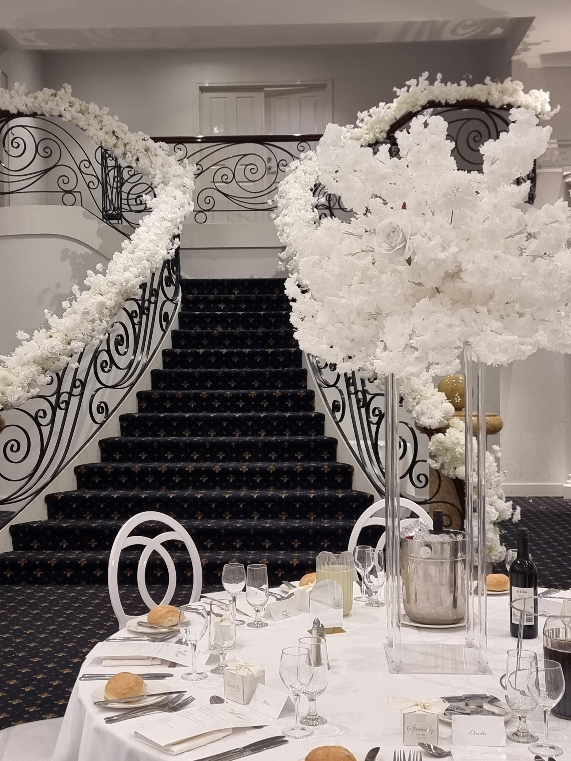 Elegant wedding reception table set beneath a grand curved staircase adorned with cascading white floral installations