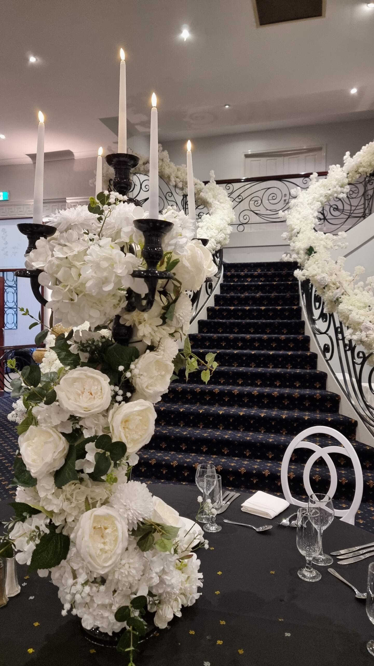 Black candelabra with white wedding florals on a reception table in front of a grand curved staircase