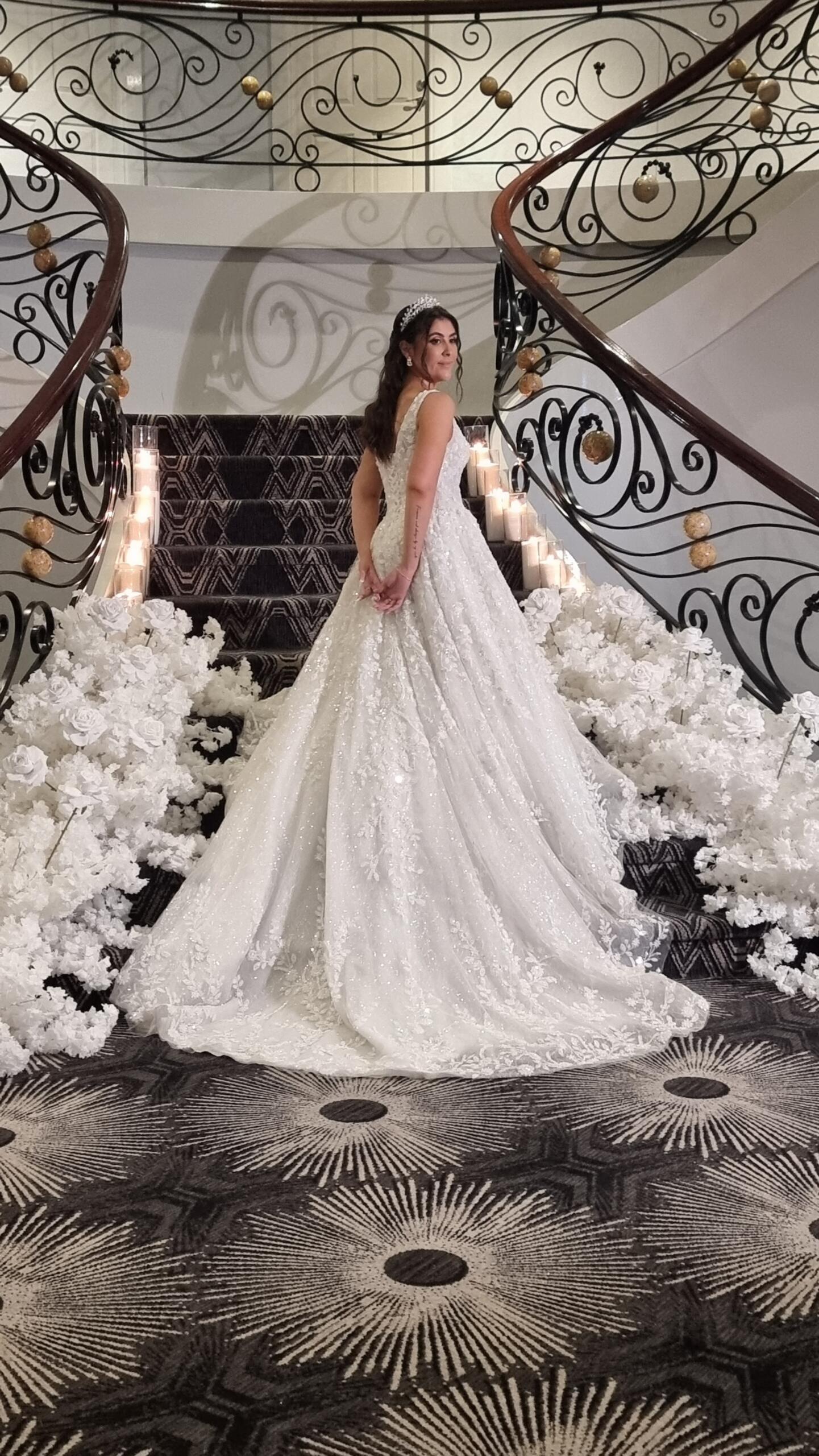 Bride in a lace wedding gown standing on a grand curved staircase lined with white florals and candlelight